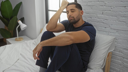 Hispanic man sitting on a bed in a stylish bedroom, reflecting with a thoughtful expression, surrounded by modern interior elements including a plant and white brick wall.