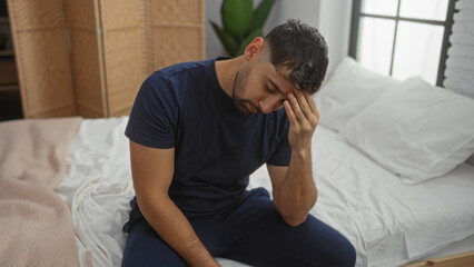 Young man sitting on bed in bedroom with thoughtful expression, hands touching head, casual attire, suggesting contemplation and introspection during morning routine indoors