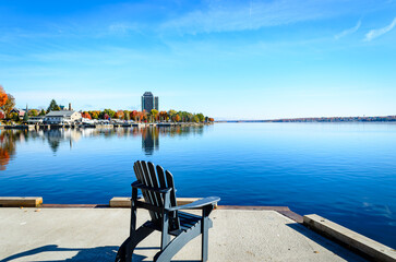 Fototapeta premium Single empty Adirondack chair on a cement pier overlooking a centered view of a shoreline marina surrounded by fall colors with blue rippling water in the foreground.