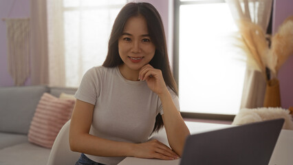 Beautiful young chinese woman sitting in a cozy home living room with soft lighting and a laptop, wearing a casual white t-shirt, smiling and looking at the camera