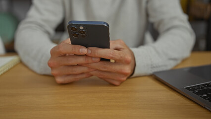Man using smartphone with both hands in modern office setting near laptop on a wooden table, portraying a tech-savvy workplace atmosphere with focus on digital communication.