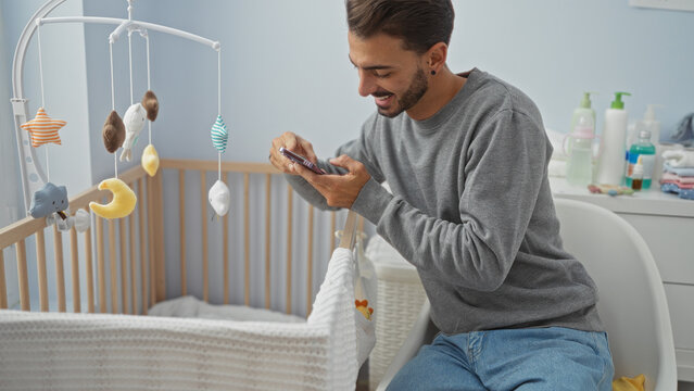 Handsome young man in bedroom taking photo of baby in cradle with smartphone, showing a heartwarming and intimate family moment indoors.