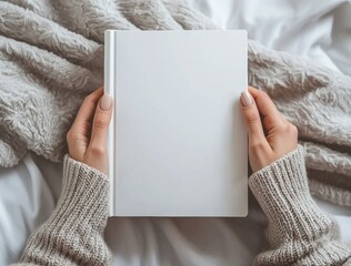 Woman wearing a comfortable sweater is holding a white blank book cover mockup while lying in bed, perfect for showcasing book cover designs