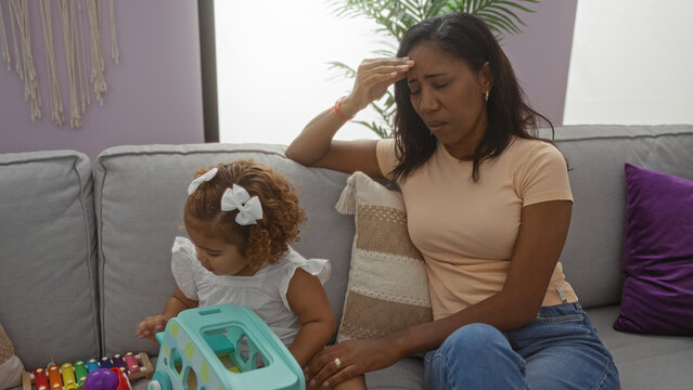 Mother sitting stressed on a sofa in living room with daughter playing nearby depicting family life indoors.