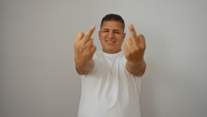 Young hispanic man wears a white t-shirt and smiles while showing middle fingers over an isolated...