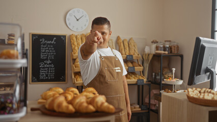 Young hispanic man working in a bakery pointing towards the camera in an indoor setting filled with different varieties of bread and pastries