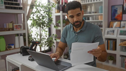 Young man working in a home decor shop, analyzing documents on a laptop amidst a modern and stylish interior