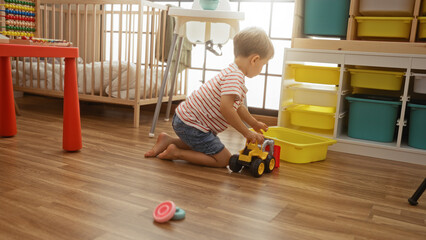 Toddler boy playing with a toy truck on the floor of a brightly lit indoor play area, surrounded by...
