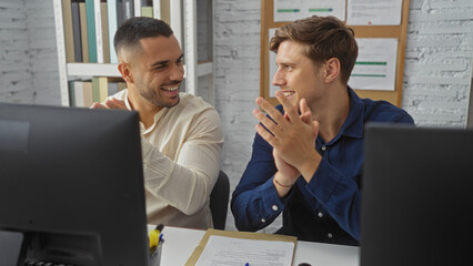 Handsome men working indoors clapping at computers signifying teamwork and happiness in a collaborative office environment.