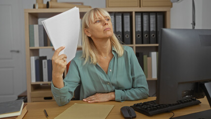 Blonde woman sitting at her desk in an office holding papers while contemplating with a serious expression in a professional indoor setting