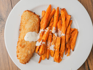 Popular fish and chips meal on a white plate and brown wooden table. Chips are made from sweet potatoes and cut in long chunks. There is splash of sous or cream on top of fine food.