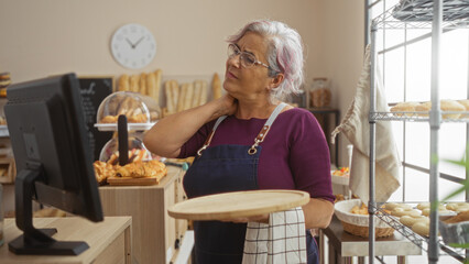 Elderly woman with grey hair in a bakery suffers from neck pain while holding a wooden tray indoors in a shop setting with bread and pastries around