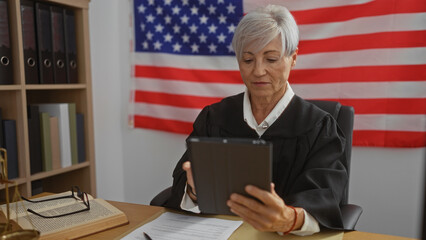 Female judge with grey hair sits in an office, reviewing documents on a tablet, with the united...