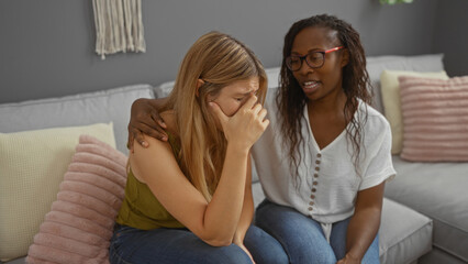 Woman comforting a crying friend sitting together on a couch in a cozy living room, displaying...