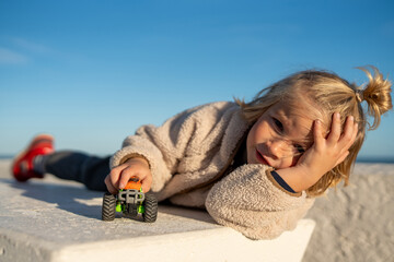 Thoughtful Child Lying on a White Concrete Surface, Playing with a Toy Monster Truck Under a Clear Blue Sky © sutulastock