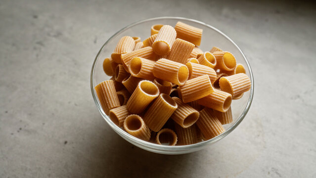 Uncooked pasta arranged neatly in a clear glass bowl against a textured grey background, highlighting its cylindrical shape and ribbed texture.
