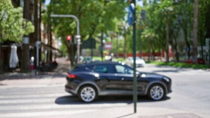Blurred black car drives along a defocused street with green trees and buildings in the bokeh background during a sunny outdoor day