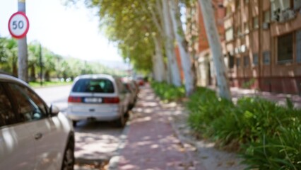 Blurry outdoor street scene with defocused cars parked along a blurry sidewalk, shaded by large leafy trees with a bokeh background effect on a sunny day