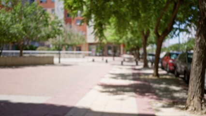 Defocused outdoor scene of a sunny urban street lined with trees, cars parked alongside and buildings in the background, creating a blurred and bokeh effect.