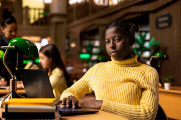 African american schoolgirl writing class notes at a study desk in a library, working hard on a school project with online information on laptop. Pupil preparing for lessons at the academic institute.