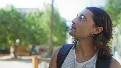 Handsome young hispanic man with long hair in an urban park looks thoughtfully into the distance