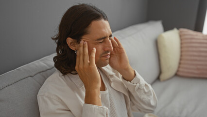Young hispanic man with long hair sitting on a sofa in a living room touching his temples and closing his eyes, indicating stress or headache, in a cozy indoor setting