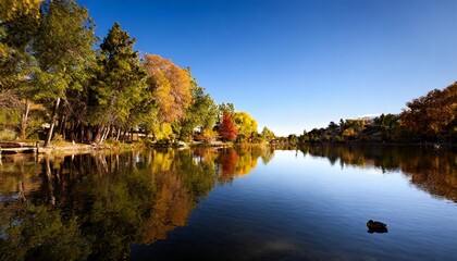 A serene lakeside view with calm water reflecting the vibrant colors of autumn trees under a clear blue sky.