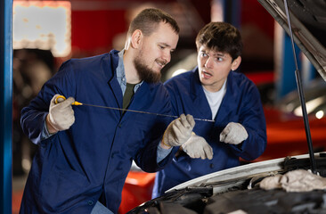 Two auto mechanics checking engine oil level under the hood of car together in a garage