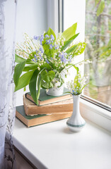 two bouquets of spring flowers on the windowsill, one of which stands on a stack of old books, lilies of the valley, creeping blue phlox,atmospheric scene,
