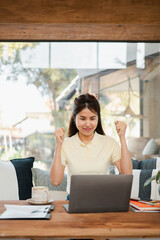 A joyful young woman in a casual setting celebrates success while working on her laptop, surrounded by a cozy home environment.