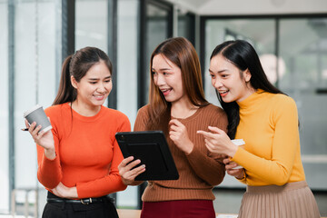 Three women in a modern office, smiling and interacting with a tablet, enjoying a coffee break together.