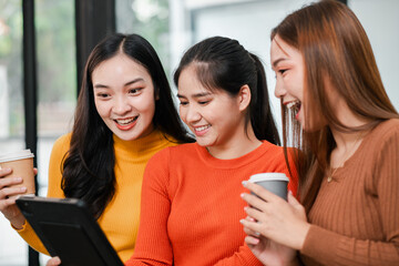 Three smiling young women in a cafe, sharing a digital tablet and enjoying coffee together.