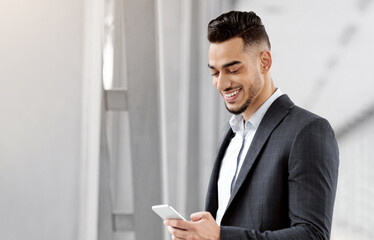 Fototapeta premium Business Trip. Handsome Young Arab Businessman Using Smartphone In Airport Terminal, Smiling Middle Eastern Entrepreneur Holding Passport With Tickets, Waiting For Flight Boarding, Free Space