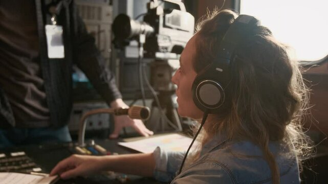 Side medium closeup of Caucasian female journalist putting on headphones, reading news report script, marking intonation, while preparing for broadcast in TV truck, chatting to operator, smiling