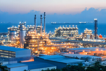Fototapeta premium Illuminated Industrial Refinery Complex Stands Tall at Dusk with Ocean Vessels in the Distance