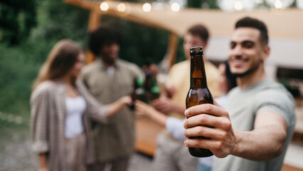 Happy young Arab guy with his diverse friends toasting with beer bottles, drinking alcohol near RV outdoors, selective focus. Millennial people enjoying summer party on camping trip, copy space