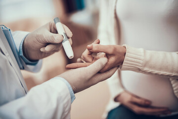 Indian doctor in white gown seeing patients in office. Doctor is measuring blood sugar of pregnant woman with daughter.