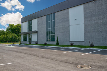 Modern commercial building with large glass windows, gray brick facade, and an empty parking lot. The structure is surrounded by greenery under a bright blue sky with scattered white clouds.