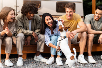 Group of multiethnic millennial friends sitting near camper van, playing with pet dog, having fun...