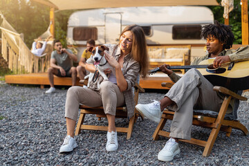 Young black guy playing guitar and singing, Caucasian lady holding cute dog near RV, camping with diverse friends in summer, having cool vacation, spending time in countryside