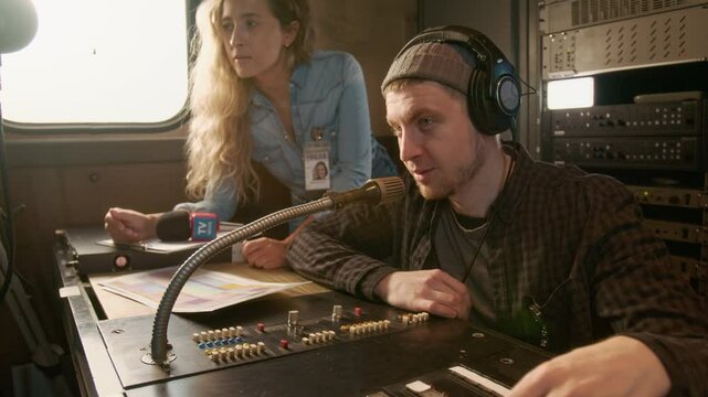 Medium shot of male TV news channel editor and female journalist with press pass sitting at workstation in production van, talking to director by video broadcast, learning details of next assignment