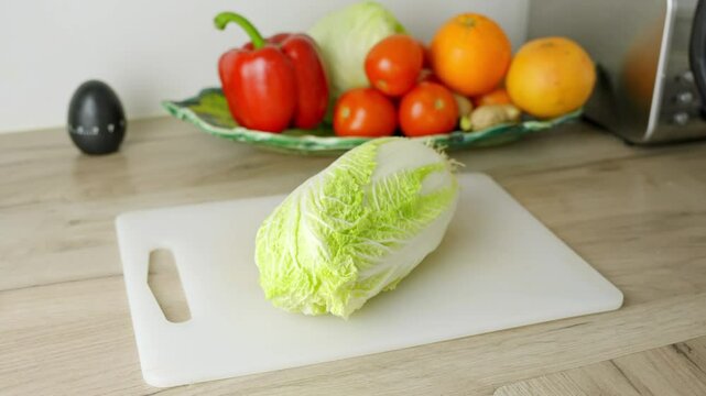 Fresh chinese lettuce round napa cabbage lying on a white cutting board in the kitchen, with other vegetables like tomatoes and peppers blurred in the background pulling out shot