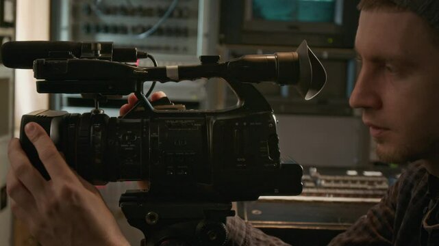 Side closeup of hands and face of busy Caucasian operator in beanie setting up video camera for filming news episode in TV production trailer, looking into viewfinder, adjusting focus and controls