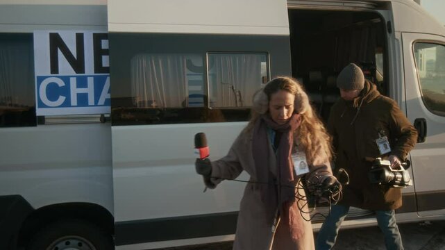 Medium full shot of Caucasian female journalist in warm coat, scarf, furry headphones and operator with camera and tripod walking out of TV production van to film news report outdoors, on winter day