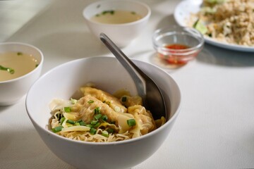 A bowl of dry egg noodles with shrimp wonton (ba mee haeng), and a plate of crab fried rice in the background, served with chicken broth at a street food stall in Silom - Bang Rak, Bangkok, Thailand
