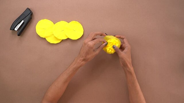 A person crafts yellow crepe paper flowers with a stapler on a brown surface