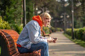 Fototapeta premium Thoughtful blond man in brightly colored outfit sits on bench in public park, anxiously waiting for date with girl, writing message on smartphone, browsing social media to pass free time.
