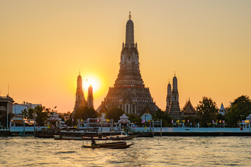 Fototapeta premium A stunning sunset envelops Wat Arun, its intricate spires silhouetted against the golden sky. Local boats softly glide along the Chao Phraya River, creating a serene atmosphere in vibrant Bangkok.