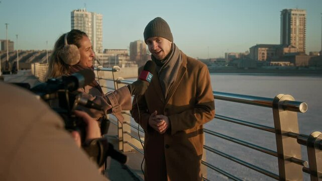 Medium full shot of unrecognizable operator with camera filming Caucasian female TV journalist interviewing male politician or city mayor in smart winter coat on occasion of opening new river bridge