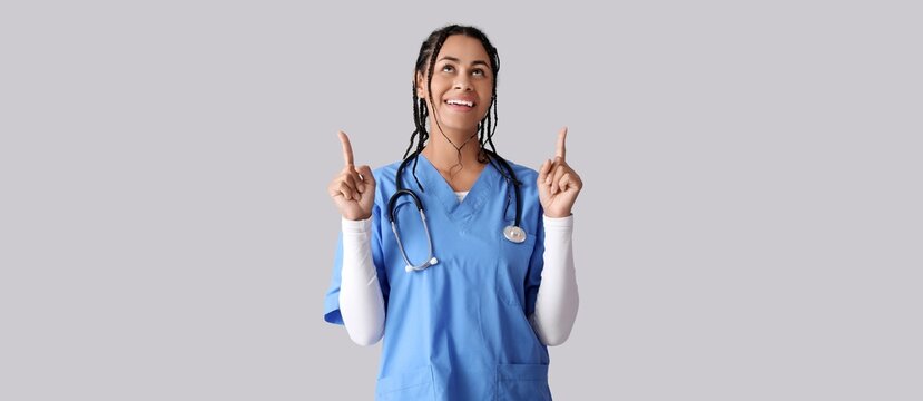 Young African-American female doctor pointing upwards on grey background with space for text